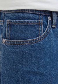 Zbliżenie na kieszeń niebieskich dżinsów z pomarańczowym przeszyciem i metalowymi nitami u osoby noszącej białą koszulę.