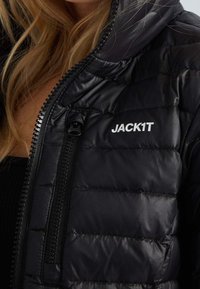 Sort puffjakke med quiltet design, forsynet med en frontlynlås og "JACK1T" logo i hvid på den øverste venstre side. Glat tekstur.