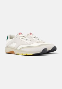Paire de baskets blanches et beiges avec des lacets blancs, des semelles jaunes et marron, des accents verts au talon et des étiquettes de logo rouges sur la languette.