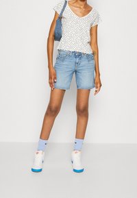 Valkoinen kukkakuosiset paita, jossa on röyhelöhihoja, vaaleansiniset farkkushortsit ja valkoiset lenkkarit sinisillä yksityiskohdilla. Yksinkertainen tausta.