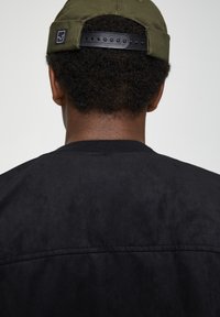 Veste bomber noire texturée avec un col côtelé ; coutures apparentes. Casquette vert olive avec une sangle noire réglable en arrière-plan.