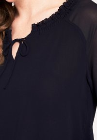Blusa blu navy con scollatura arricciata, dotata di un dettaglio con fiocco sul davanti. Tessuto trasparente con una consistenza morbida, drappeggio sottile.