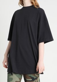 Př oversized černé bavlněné tričko s kulatým výstřihem a krátkými rukávy, nošené s kalhotami s kamuflážním vzorem. Hladká textura, volný střih.