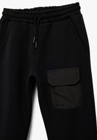 Pantalones jogger negros hechos de tela suave, con cinturilla elástica, cordón ajustable, bolsillos laterales y un bolsillo de parche en el muslo.