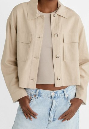 Personne portant une veste courte beige à boutons sur un crop top côtelé beige et un jean taille haute bleu clair, les mains dans les poches.