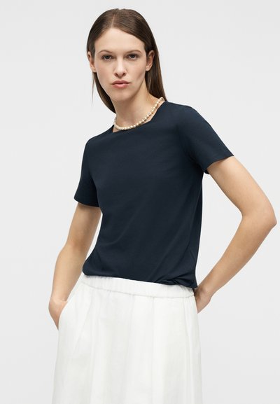 Γυναίκα με μπλε μπλούζα με κοντά μανίκια, λευκή φούστα και πέρλες στο λαιμό, στέκεται με τα χέρια στις τσέπες μπροστά σε απλό φόντο.