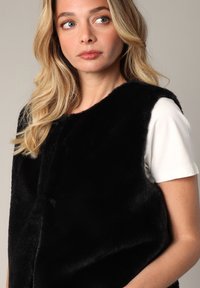 Zwarte nepbont gilet met een eenvoudige ronde halslijn, zachte textuur en geen zichtbare sluitingen, gedragen over een wit T-shirt met korte mouwen.