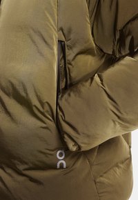 Olivgrüne Latzjacke mit einer glänzenden, glatten Textur. Verfügt über eine Reißverschlusstasche und ein Logo auf der unteren linken Seite.