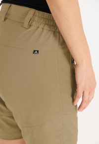 Khaki shorts dengan pinggang elastis, dilengkapi dengan kantong zipper samping dan label logo hitam kecil di bagian belakang dekat pinggang.