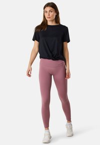 Sort kortærmet top med knude-detalje, parret med tætsiddende blush pink leggings. Hvide træningssko fuldender outfittet.