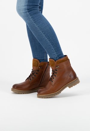 Personne portant un jean bleu et des bottines en cuir marron à lacets avec une fermeture éclair latérale et une semelle épaisse, debout sur fond blanc.