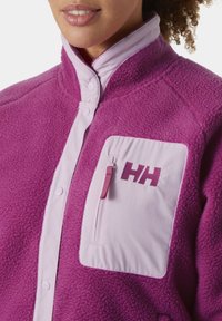 Paarse fleece jack met een hoge kraag, roze accenten en een zipzak met een logo. Zachte textuur met knoopsluiting.