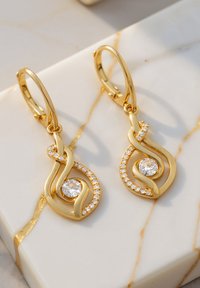 Guldtonade örhängen med svängande mönster, en central rund klar ädelsten och omgivande små klara stenar längs sidorna.