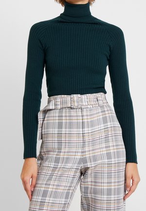 Femme portant un pull à col roulé côtelé vert foncé, rentré dans un pantalon taille haute à carreaux blancs avec une ceinture assortie.
