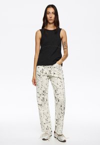 Top negro de canalé sin mangas con tachuelas plateadas en el dobladillo, combinado con pantalones en color blanco roto que presentan un patrón de salpicaduras negras y detalles de bolsillos.