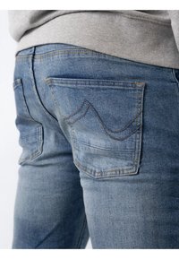 Jeans din denim de culoare albastru deschis, prezentând două buzunare spate cu un design distinctiv de cusătură ondulată și o textură netedă.