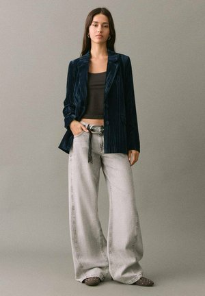 Blazer in velluto a righe navy su un crop top nero, abbinato a jeans a gamba larga grigio chiaro e scarpe marroni con dettagli bianchi. Accessori semplici.