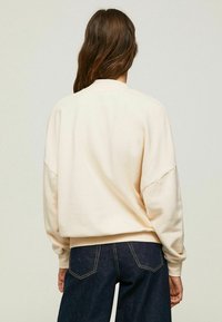 Felpa color crema con spalle scese e orlo arrotondato, abbinata a jeans in denim scuro con cuciture a contrasto.