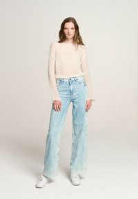 Maglione leggero e trasparente di colore crema con maniche lunghe, abbinato a jeans azzurri a gamba larga. Sneakers bianche completano il look.
