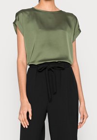 Satin olivgrüne Bluse mit aufgerollten Ärmeln, rundem Ausschnitt, kombiniert mit hochtaillierter schwarzer Hose mit gebundenem Taillenbund.