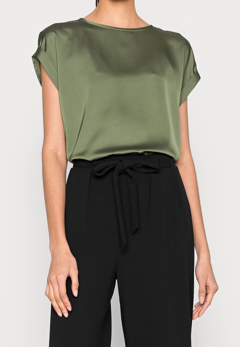 Satin olivgrüne Bluse mit aufgerollten Ärmeln, rundem Ausschnitt, kombiniert mit hochtaillierter schwarzer Hose mit gebundenem Taillenbund.