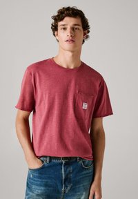 Camiseta de algodón roja con cuello redondo, mangas cortas y un bolsillo frontal que presenta una etiqueta. Llevada con jeans azules, mostrando un ajuste casual.