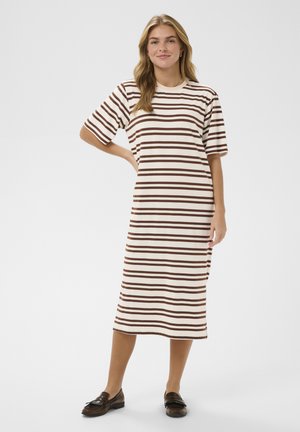 Femme debout portant une robe midi beige à rayures horizontales marron et des mocassins marron foncé, souriante avec une main sur la hanche.