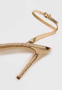 Χρυσό μεταλλικό σανδάλι με ψηλό τακούνι, υφή φιδίσιου δέρματος, ανοιχτή μύτη,  λεπτό λουράκι στο αστράγαλο και αιχμηρό, μυτερό τακούνι.