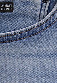Le tissu en denim bleu clair présente une texture lisse avec des coutures visibles. Une étiquette noire affiche "MAVI PRO-SPORT". Le design possède un col arrondi.