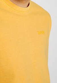 Žluté bavlněné tričko s kulatým výstřihem a malým vyšitým logem na hrudi. Hladká textura s volným střihem.