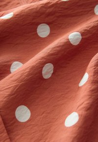 Textil mit rustfarbenem Grund und großen, weißen Punkten. Der Stoff wirkt leicht und leicht zerknittert, was zur Struktur beiträgt.