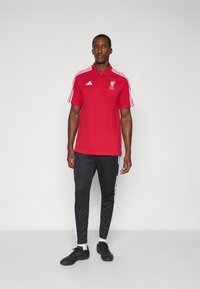 Punainen Liverpool FC:n polo-paita lyhyillä hihoilla, valkoiset raidat olkapäillä ja logo. Yhdistetty mustiin kapeneviin housuihin, joissa on sivuraidat.