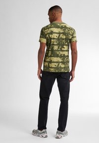 T-shirt s krátkým rukávem olivově zelené barvy s pruhovaným vzorem palmových listů, doplněný černými džíny a vícebarevnými teniskami.