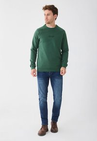 Grønn genser med rund hals og svart tekst, kombinert med blå jeans og brune støvler. Glatt stoff, avslappet passform og ribbestrikkede mansjetter.