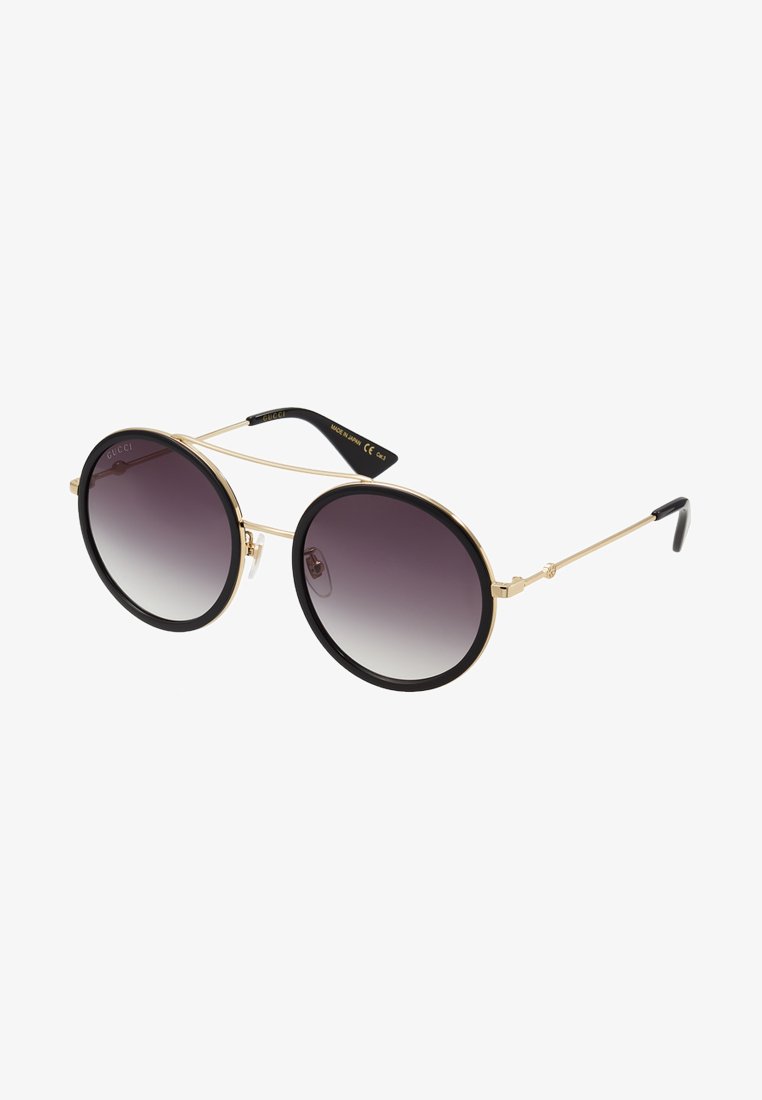Óculos de sol redondos com uma armação em metal dourado, detalhes em preto e lentes cinzas degradê. Design minimalista com hastes finas e detalhes do logo.