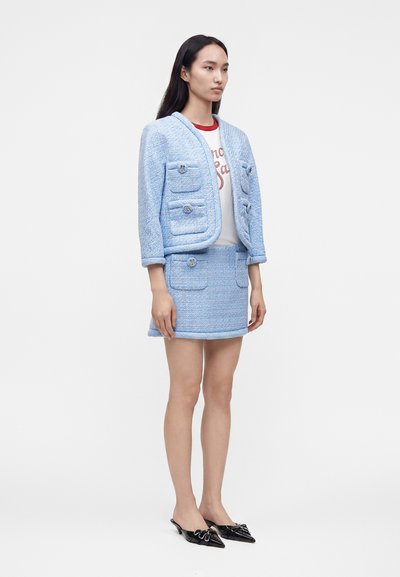 Femme debout portant un tailleur jupe deux pièces texturé bleu clair avec poches à rabat et un t-shirt blanc graphique, associé à des chaussures noires à bout pointu.