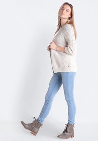 Cárdigan de punto beige con un diseño texturizado, mangas enrolladas y ventilaciones laterales, combinado con jeans ajustados azul claro y botas de tobillo grises.