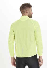 Lätt, fluorescerande gul idrottsjacka med långa ärmar, reflekterande detaljer och hög krage. Slät textur med en figurnära design.