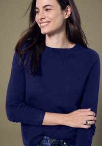 Marineblauer Pullover mit Rundhalsausschnitt, langen Ärmeln und weichem Material. Kombiniert mit Jeans und einem silbernen Ring am Handgelenk.