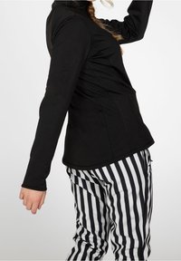 Person i en svart långärmad tröja och svarta och vita vertikala randiga byxor, som höjer ena armen mot en enkel ljus bakgrund.