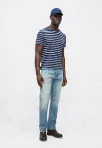 Navy plava majica na pruge, svijetloplave poderane traperice i crne kožne čizme; kačket s logotipom, minimalni detalji, ležeran kroj.