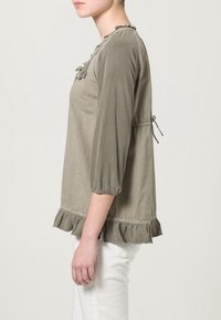 Blusa verde oliva con maniche a 3/4, orlo arricciato e dettagli sul colletto, dotata di lacci laterali regolabili, realizzata in tessuto morbido e testurizzato.
