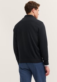 Mann in schwarzem Langarmpullover und blauen Hosen von hinten vor einem einfarbigen hellen Hintergrund gezeigt.