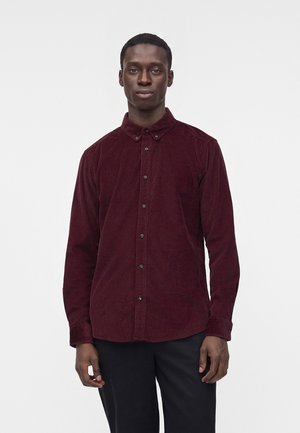Homme debout portant une chemise rouge foncé à manches longues boutonnée et un pantalon noir sur un fond blanc uni.