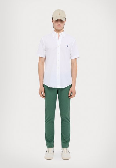 Chemise blanche à manches courtes avec col, pantalon vert et casquette beige avec un logo. Les chaussures sont de couleur claire avec une accentuation foncée.