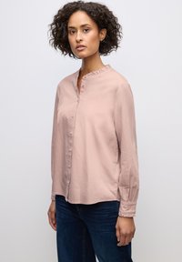 Světle růžová košile s dlouhým rukávem, s knoflíkovým zapínáním v přední části, volánkovým límcem a elastickými manžetami, vyrobená z hladkého materiálu, spárovaná s tmavě modrými džíny.