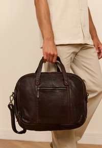 Brown leather messenger bag with two top handles, adjustable shoulder strap, and front zipper pocket. Smooth texture and structured shape.