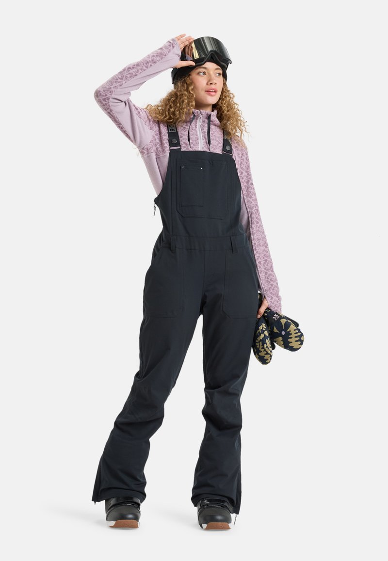Czarne kombinezony narciarskie z regulowanymi paskami, fioletowy, teksturowany longsleeve pod spodem oraz gogle narciarskie. Trzymają wzorzyste rękawiczki.