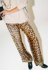 Pantaloni con stampa leopardata in un tessuto morbido e lucido, dalla vestibilità ampia, caratterizzati da macchie nere e marroni vivaci su uno sfondo beige chiaro.