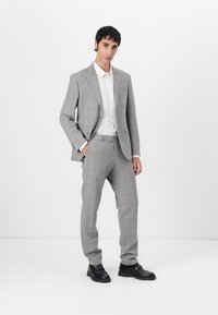 Completo grigio chiaro con un sottile motivo a check, abbinato a una camicia bianca con bottoni. Tessuto morbido, vestibilità su misura e scarpe nere.